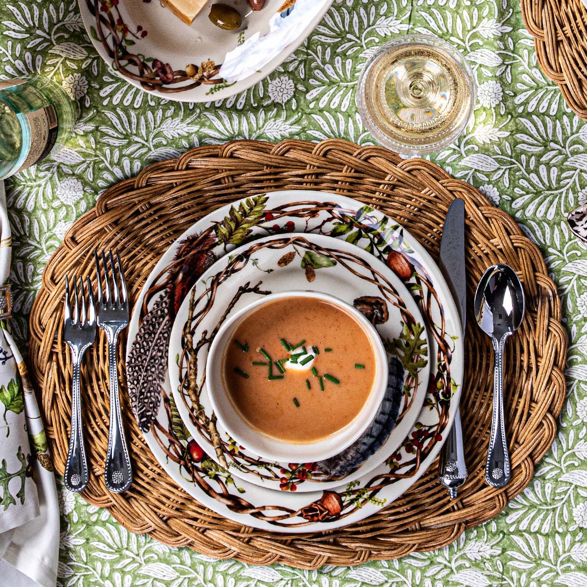 Overhead of a Forest Walk place setting with a bowl of soup on a wicker charger and green leaf tablecloth