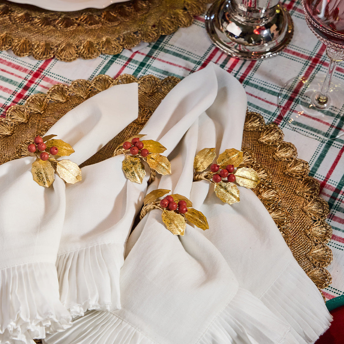 Gold holly napkin rings with red berries on folded white napkins over a plaid tablecloth