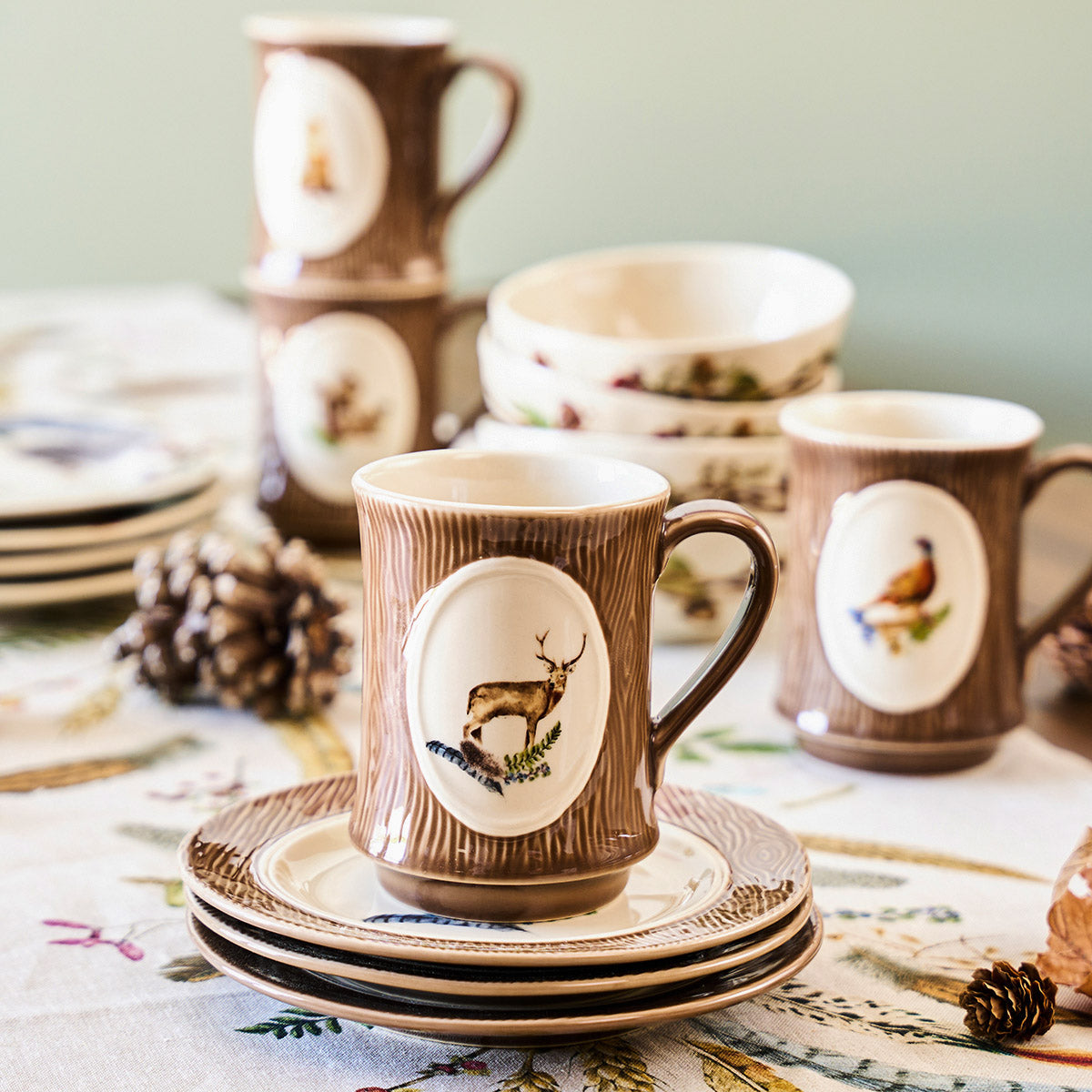 Forest Walk stag mug on stacked saucers with more animal mugs and pinecones on a patterned tablecloth