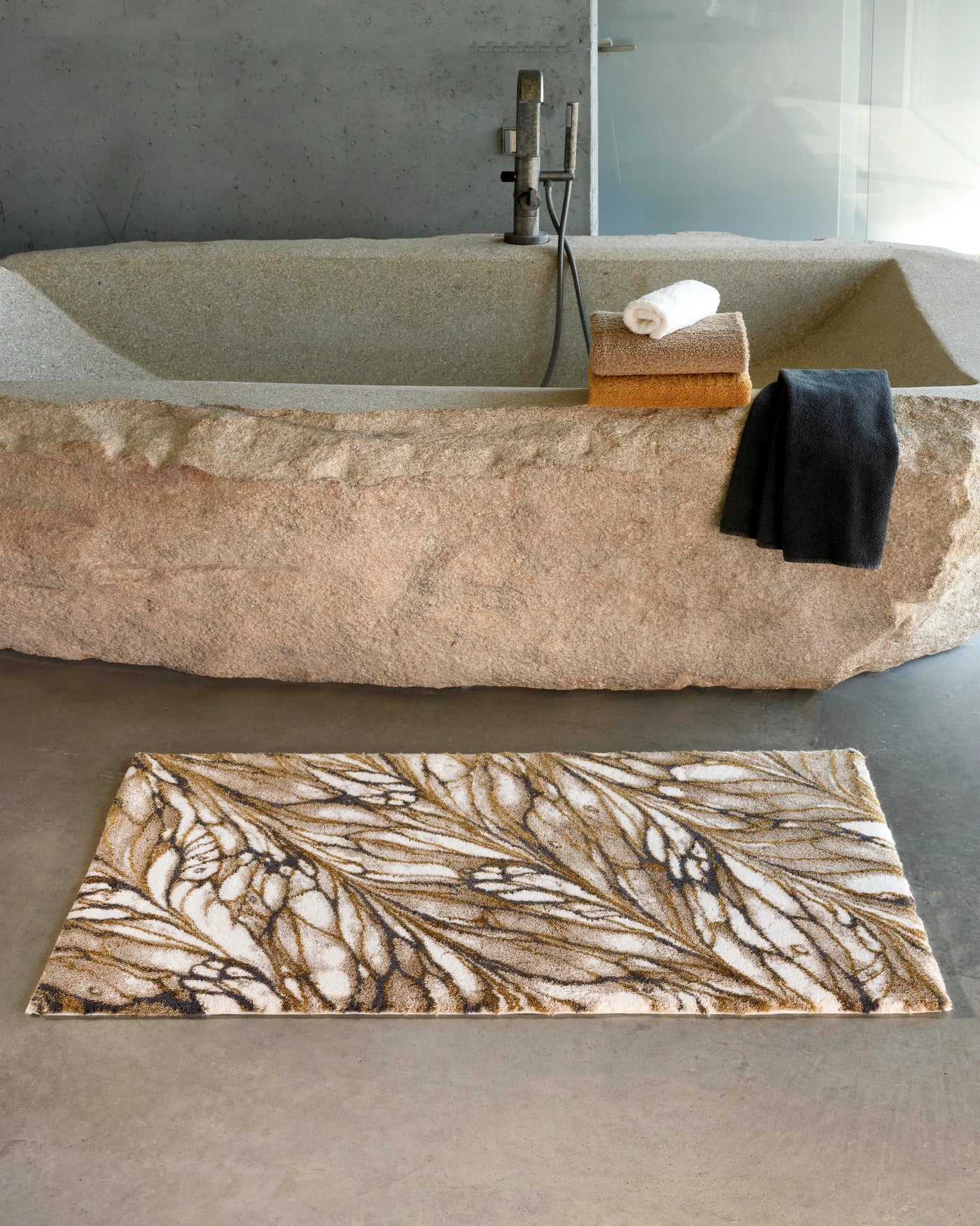 Abyss Habidecor Diane bath rug in ivory and gold botanical pattern beside stone tub.
