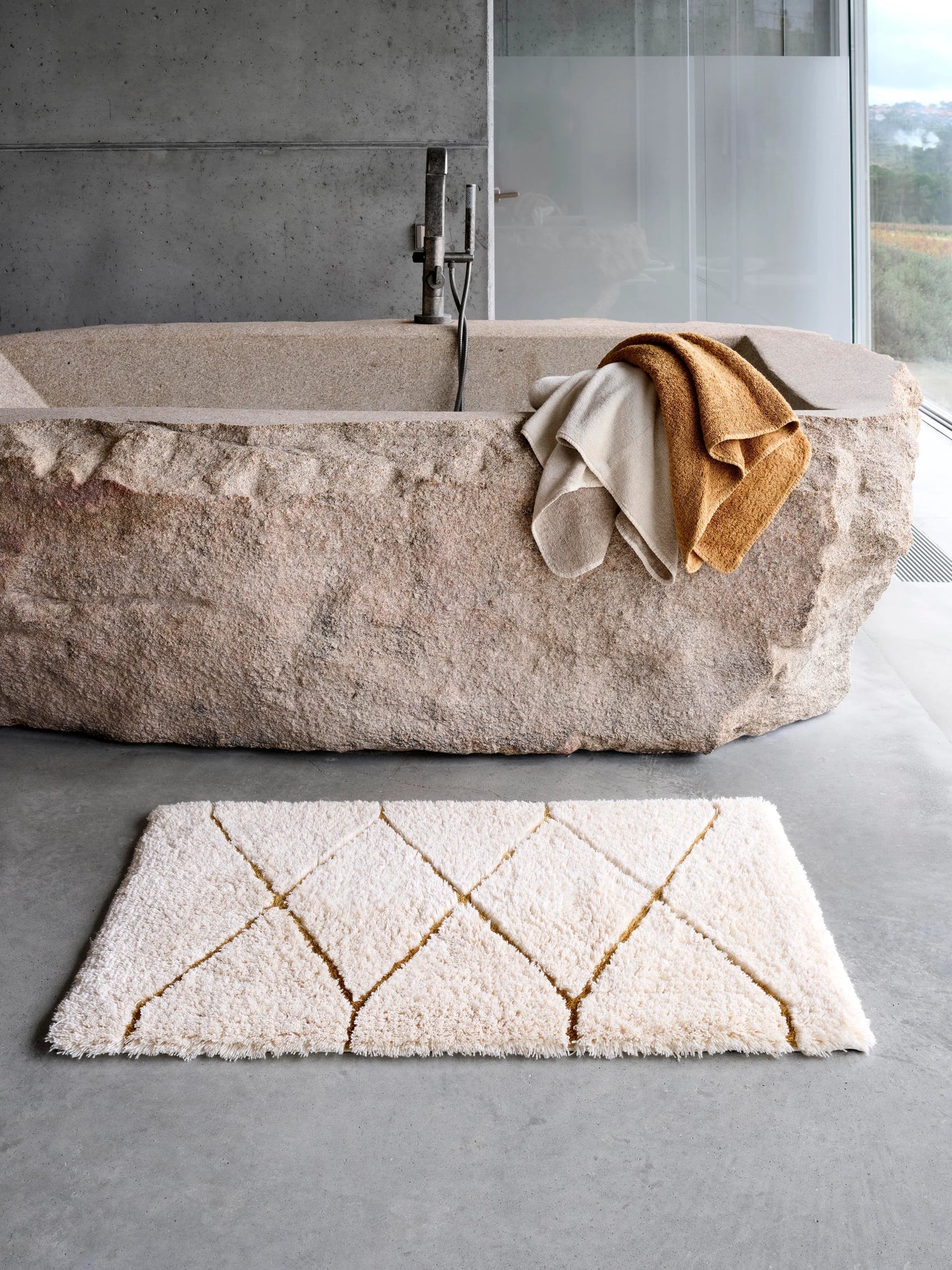 Ivory tufted bath rug with gold diamond pattern on concrete floor beside stone bathtub.