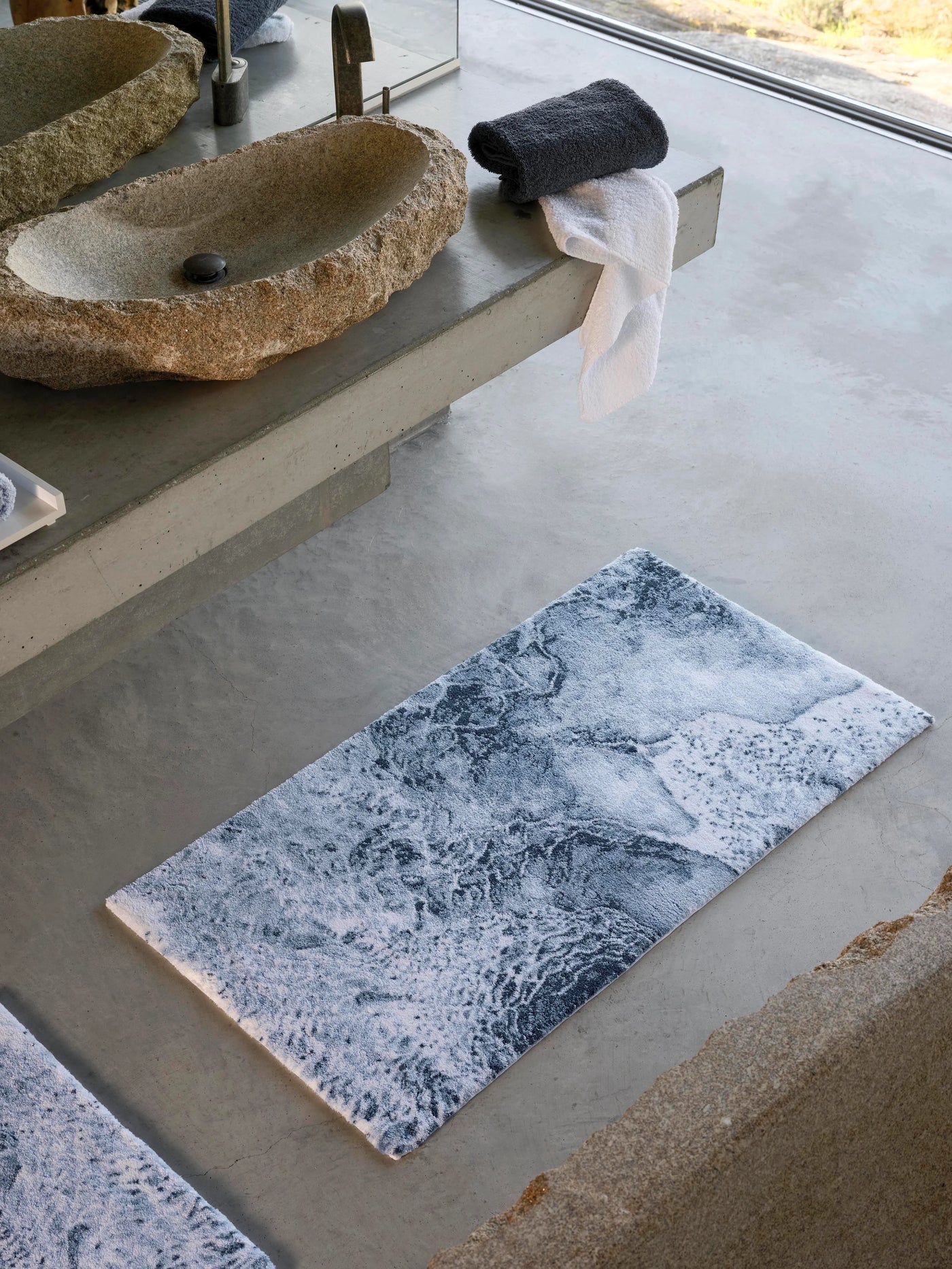 Overhead view of Nazare bath rug with ocean-wave print in slate and white on concrete floor.