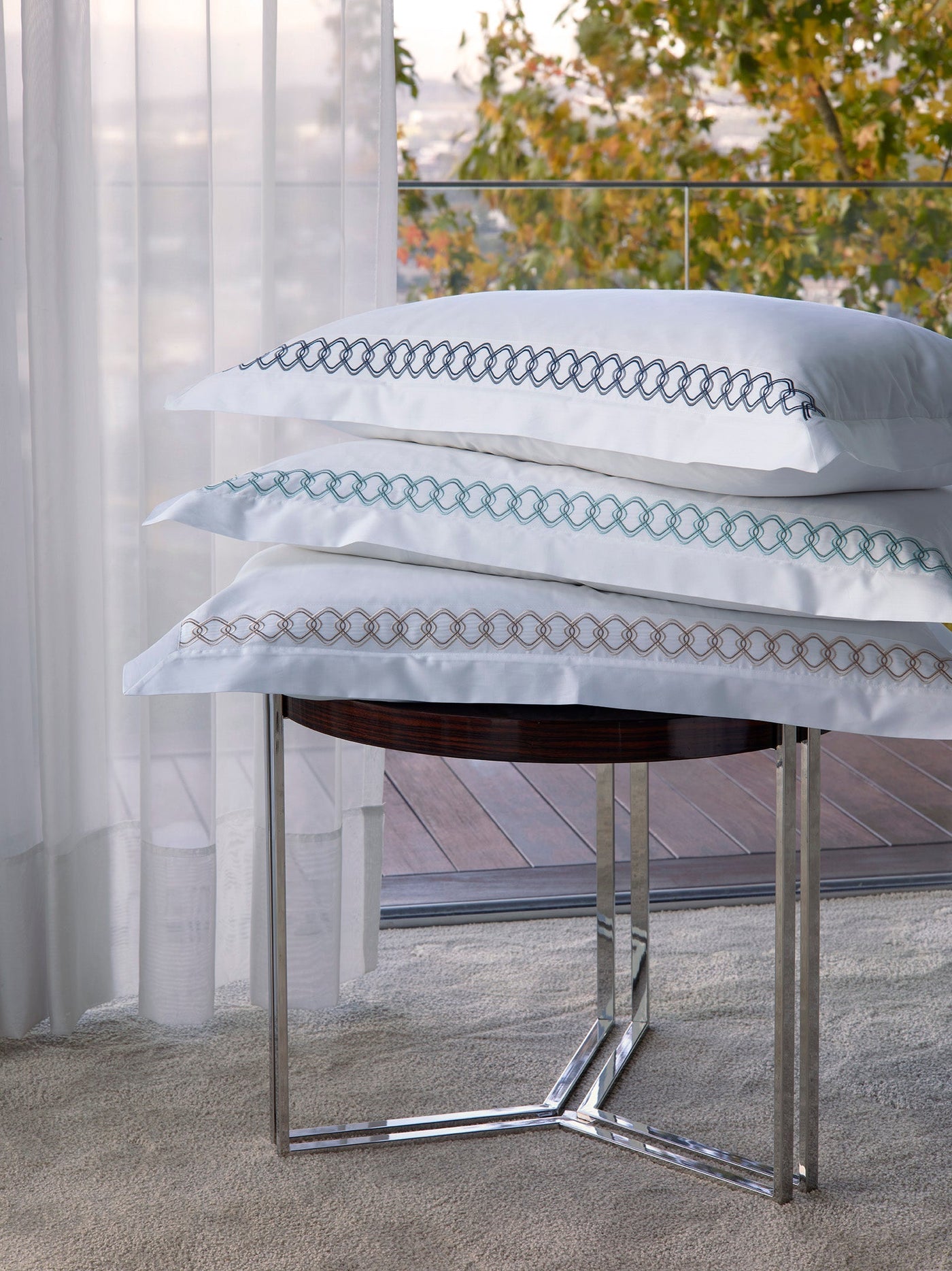 Close-up of stacked Ornato shams showing teal and taupe embroidered chain border on white cotton.