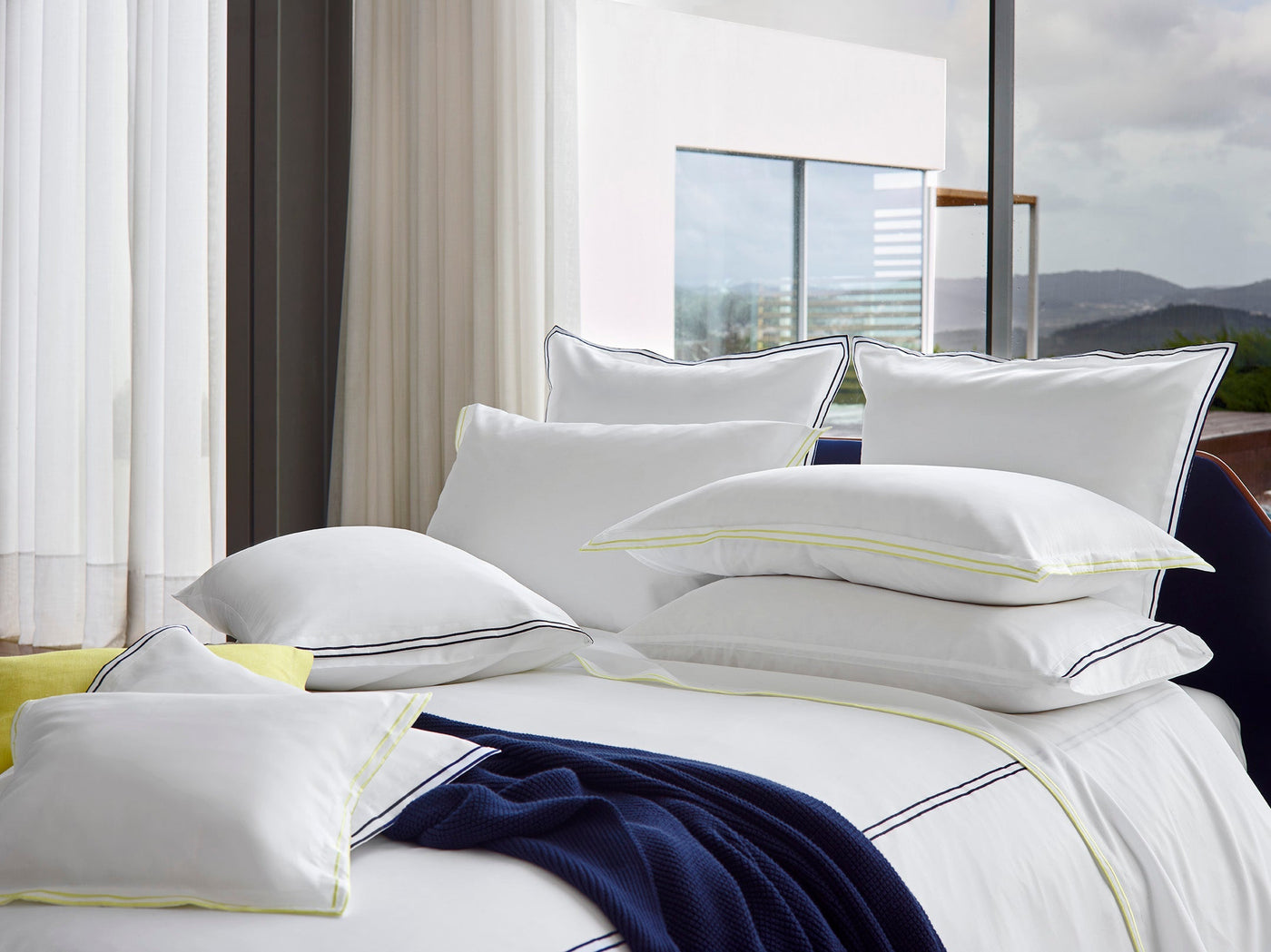 A neatly made bed features Sferra’s Sereno | Pillowcase, Pair in crisp white, accented with thin piping, beside large glass windows. A navy blue blanket lies at the foot of the bed, completing the serene look.