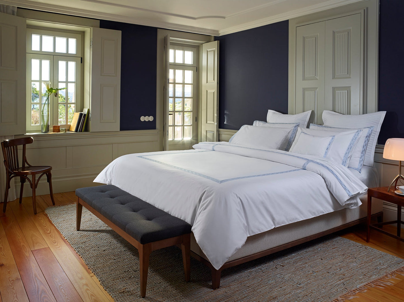 A neatly made bed with the Memoria Duvet Cover sits in a cozy bedroom featuring dark blue walls, wooden floors, a woven rug, wooden furniture, and two large windows letting in natural light.