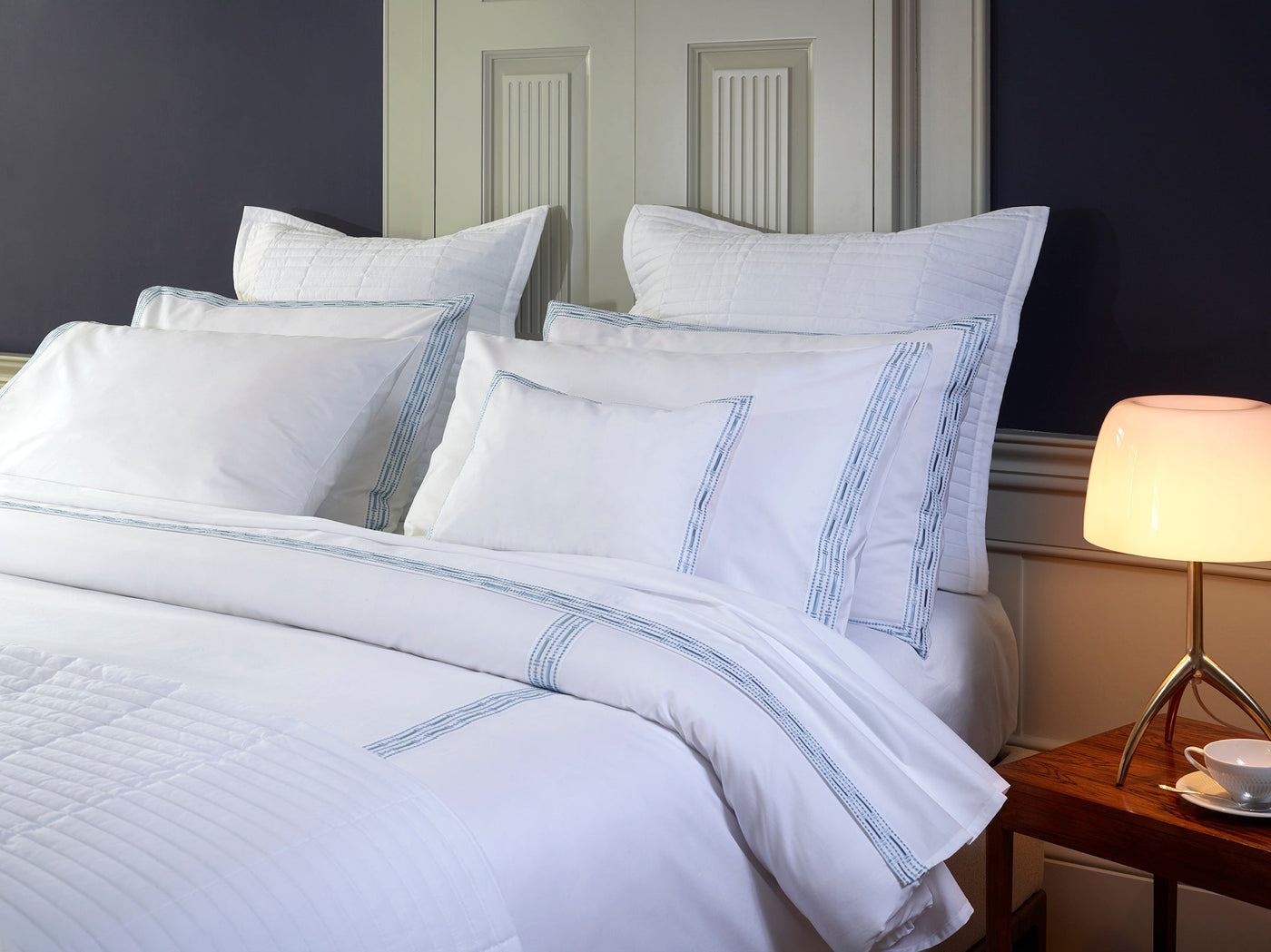 A neatly made bed with white linens and Memoria Pillowcase Pair, featuring subtle blue stripes, against a paneled wall. A modern lamp with a round white shade sits on a wooden bedside table.
