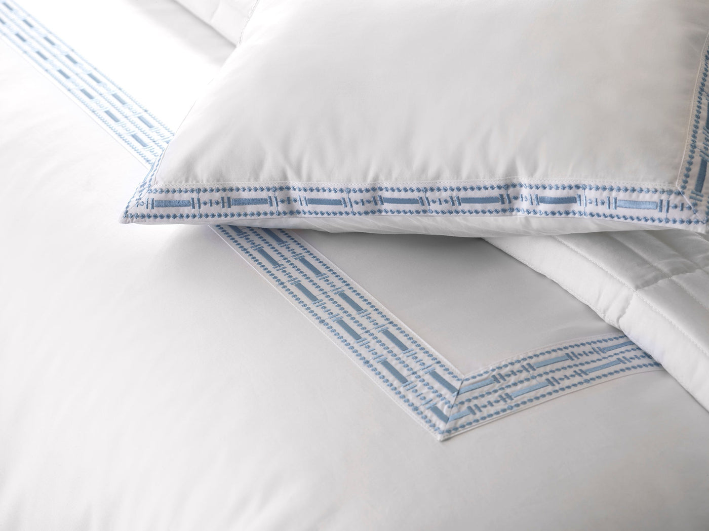 Close-up of the Memoria Duvet Cover, crafted in Portugal, displaying white bedding with a light blue geometric embroidered border and a matching pillow and sheet trim.