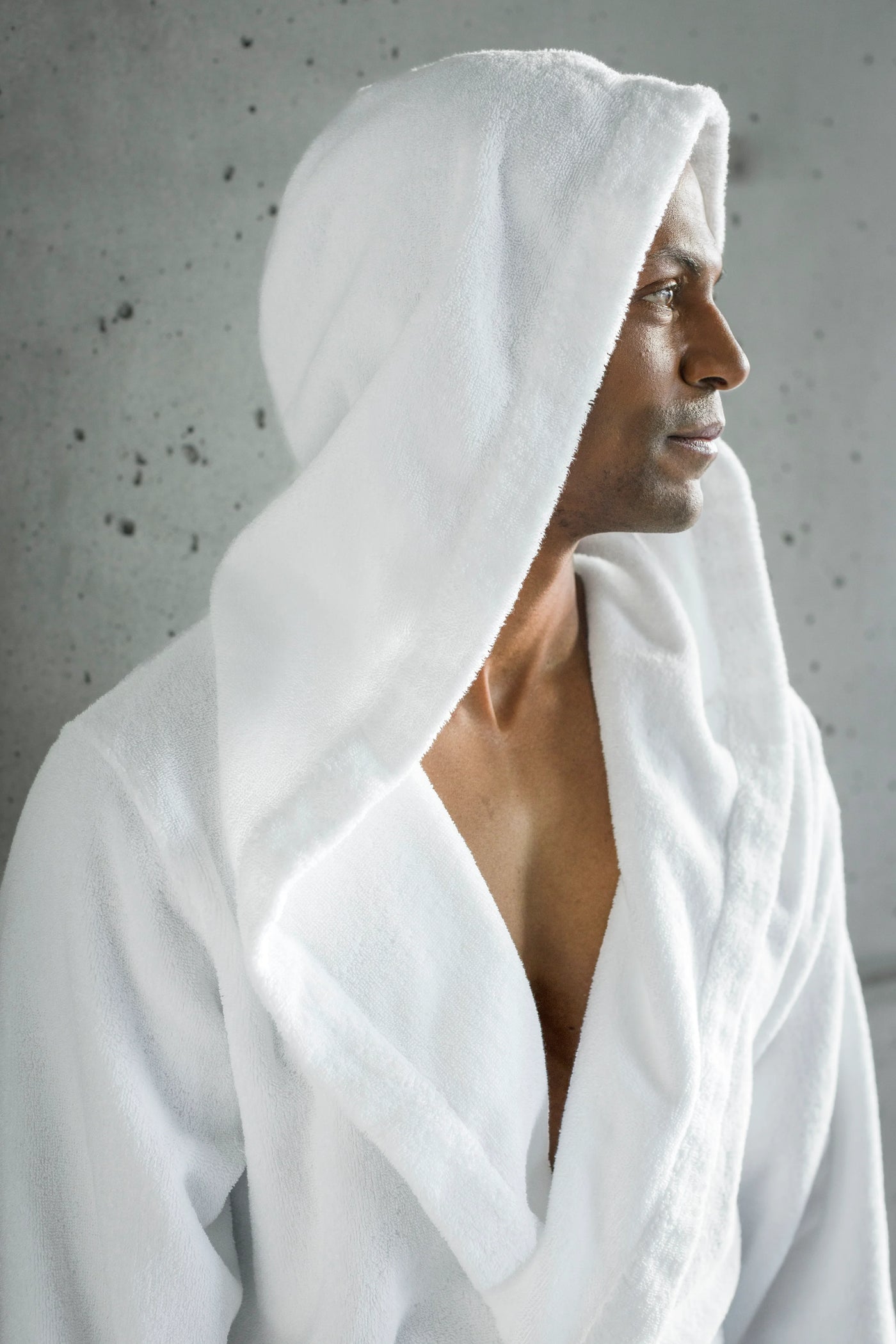 Man wearing white Abyss Habidecor hooded terry robe, profile view against concrete wall.