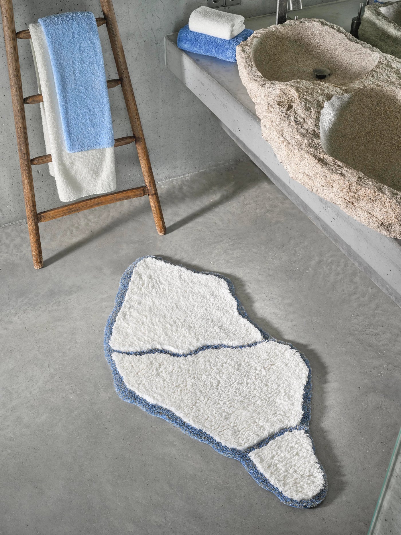 Organic-shaped white bath rug with periwinkle border on grey concrete floor.