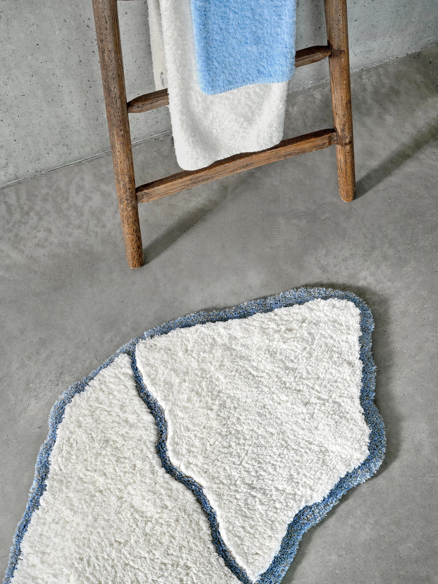 Organic-shaped white bath rug with blue wavy border, styled on concrete floor beside wooden ladder.