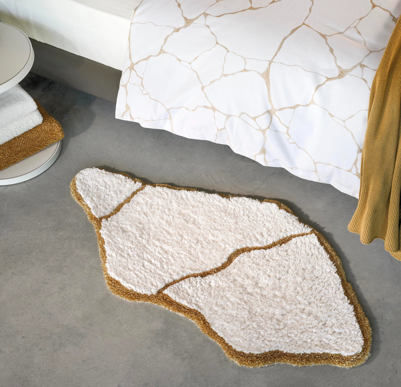 Abyss Habidecor Jess bath rug in ivory with gold border, organic stone shape on gray floor.