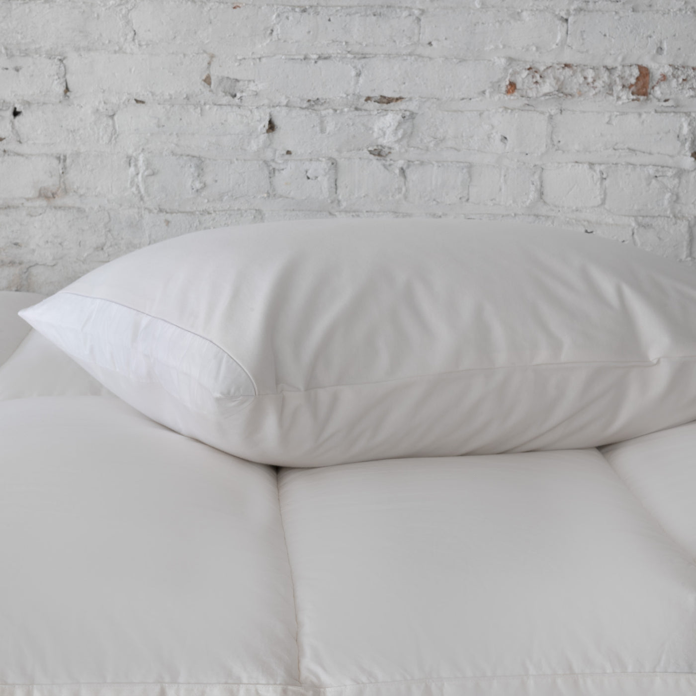 The St. Geneve Absolute | Pillow Protector in white covers a pillow placed on a white mattress topper, set against a white brick wall.