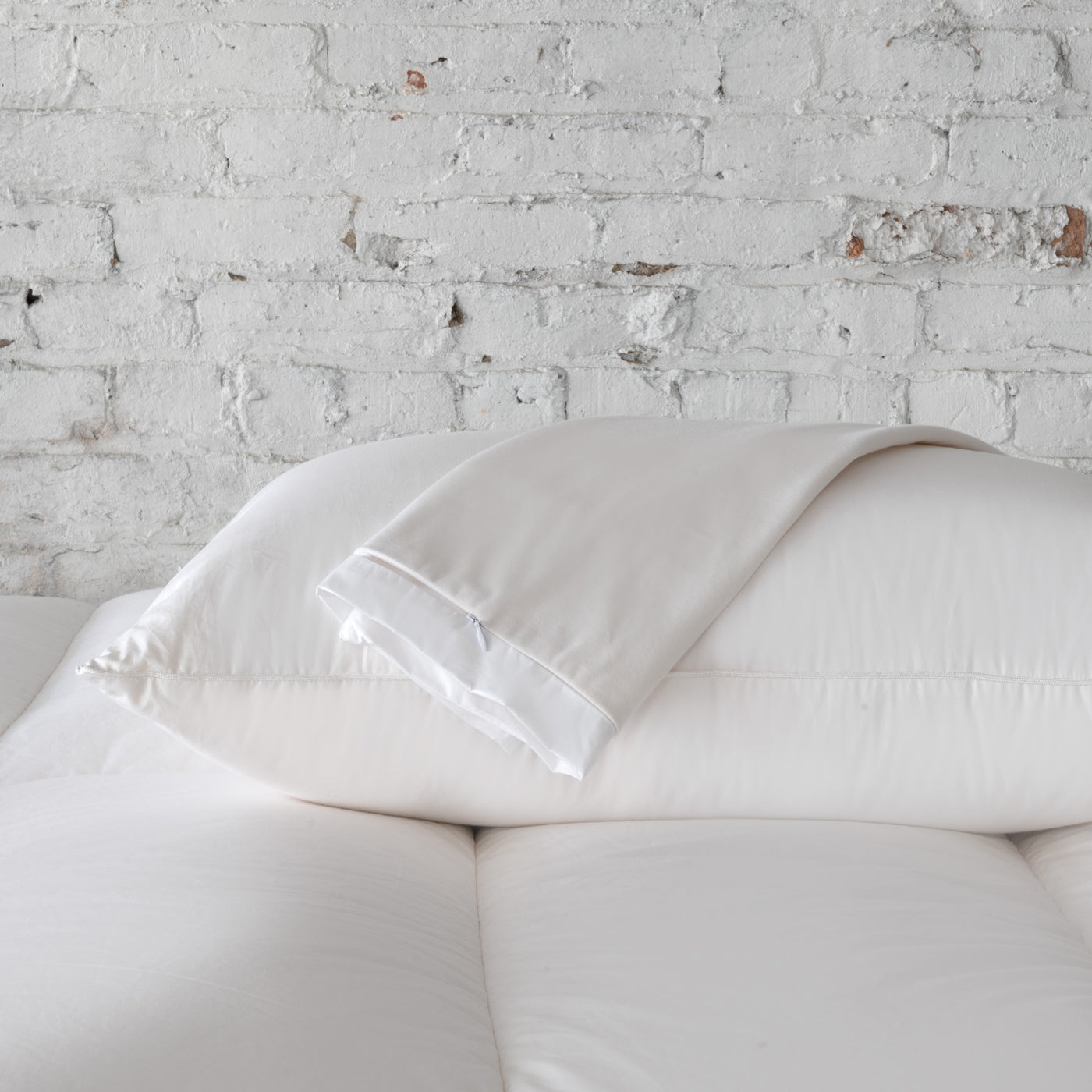 A St. Geneve Absolute | Pillow Protector is folded on top of a white pillow, which rests on a white mattress against a white brick wall background.