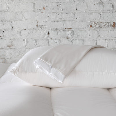 A St. Geneve Absolute | Pillow Protector is folded on top of a white pillow, which rests on a white mattress against a white brick wall background.