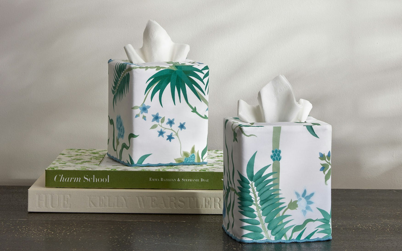 Two Matouk Palmyra tissue box covers with a green botanical pattern sit on a dark surface, with one resting atop a stack of decorative books.