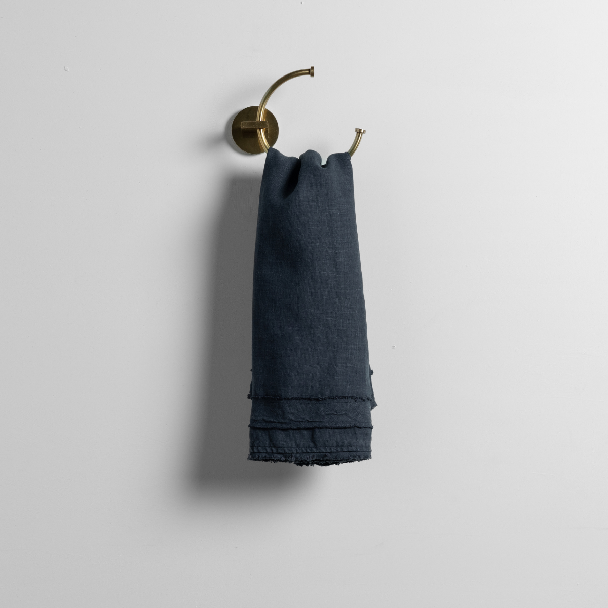 Slate linen guest towel with fringe hem, hung on a brass ring against a white wall.