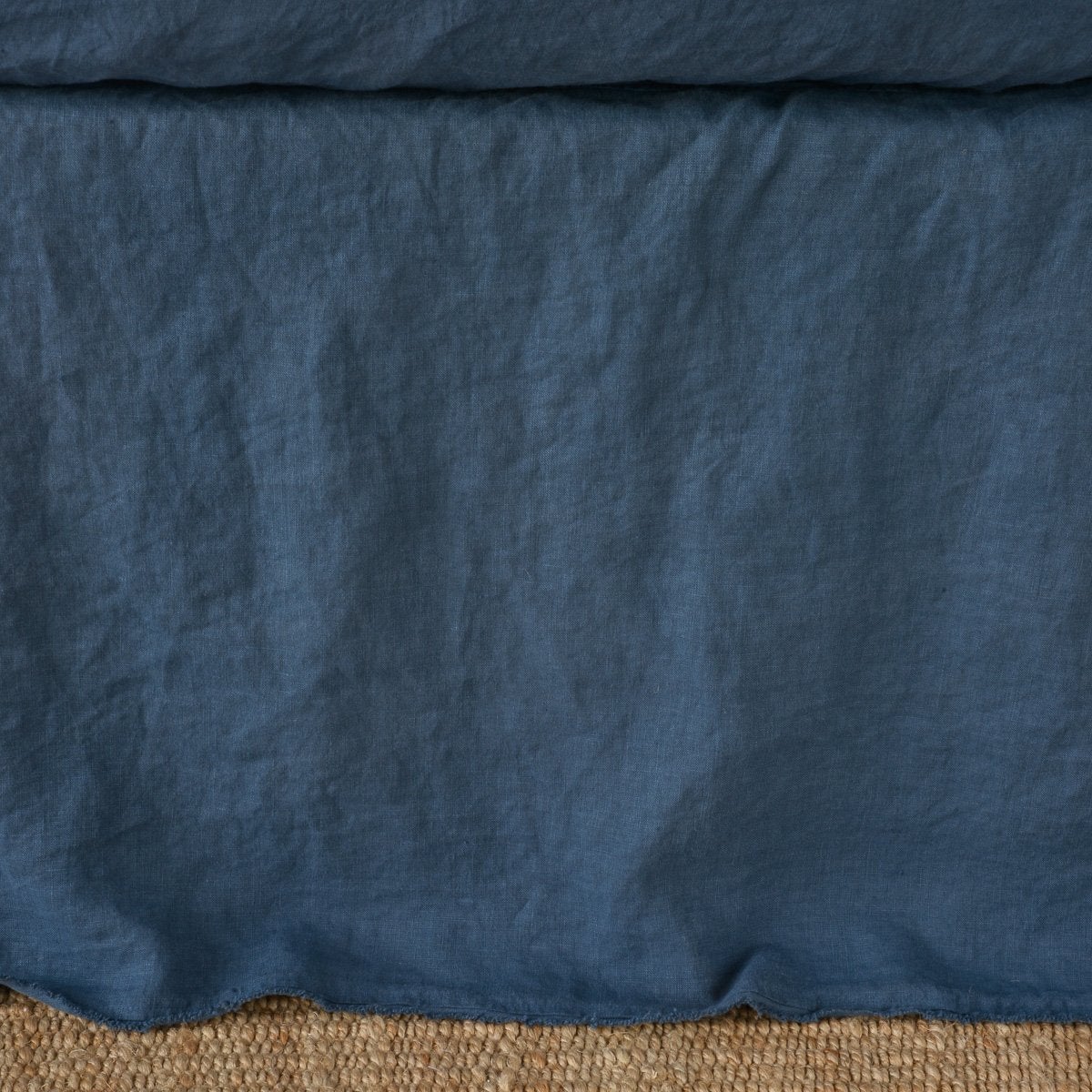 Midnight linen bed skirt with relaxed drape, resting on natural woven rug.