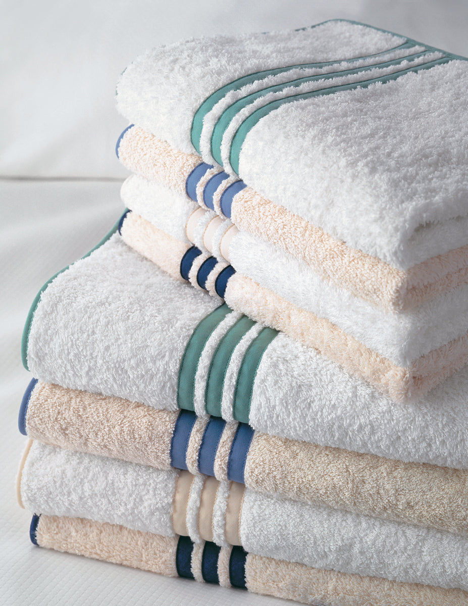 A neatly stacked pile of Matouk Newport wash cloths with green and blue stripe accents and an appliqué-tape border, arranged on a white surface.