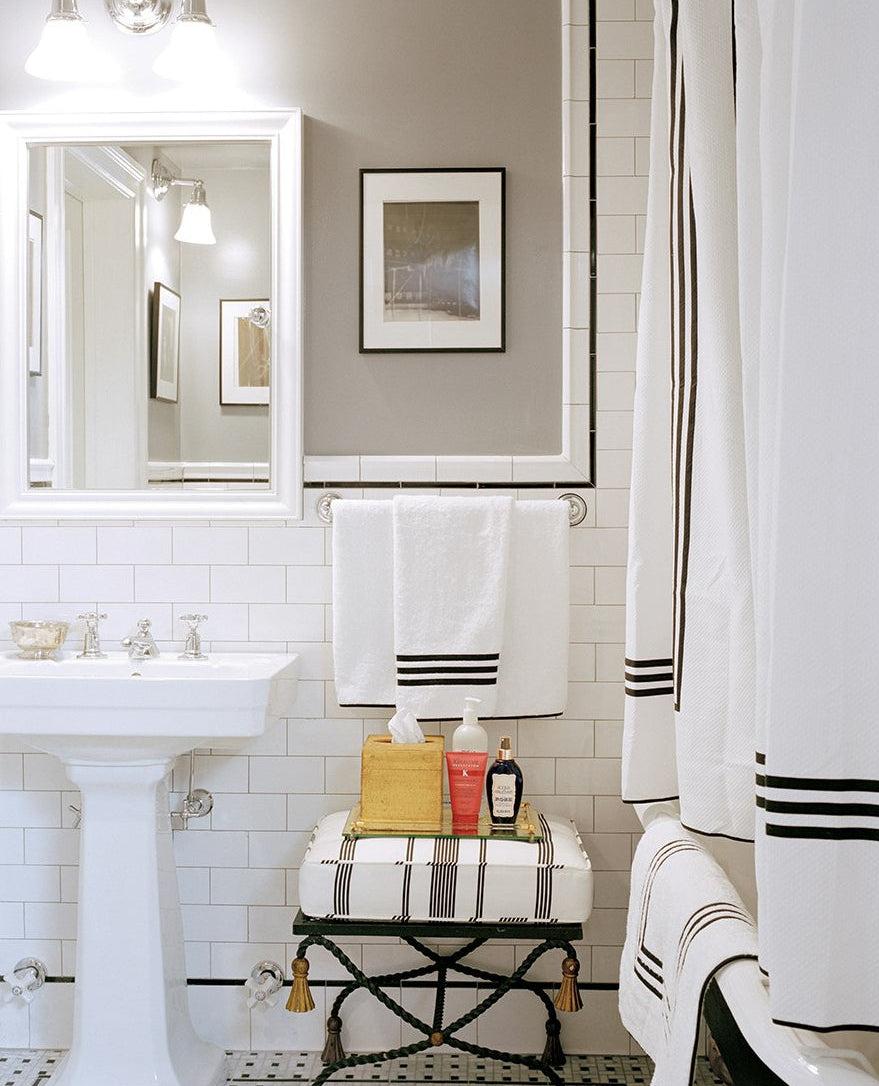 A white-tiled bathroom with a pedestal sink, mirror, towel rack with bath linens, framed photo, and stool with toiletries features the Matouk Newport Shower Curtain in elegant diamond pique and black trim.