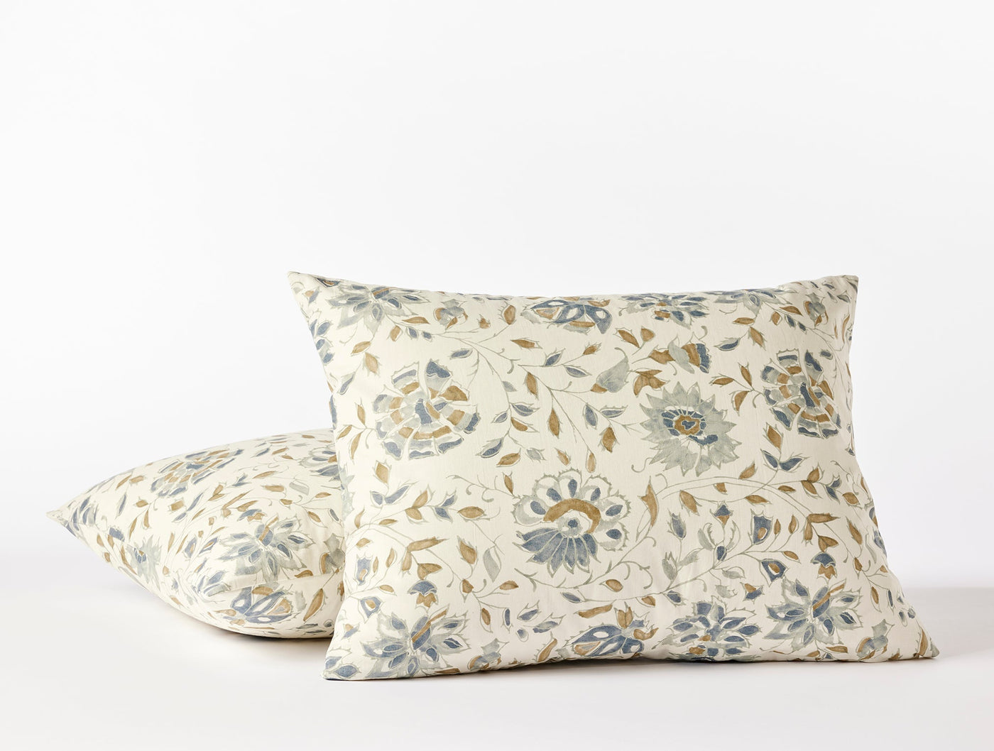 Two Coyuchi Flora Print shams in ivory with blue and gold floral motifs, stacked at an angle.