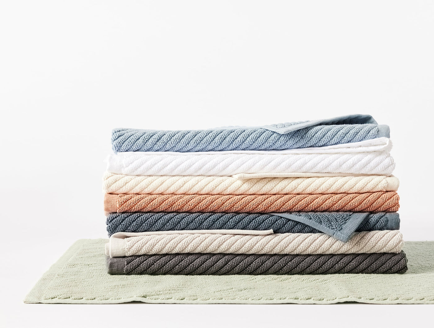 Close-up of stacked Coyuchi Air Weight bath rugs in multiple colorways on a sage green rug.