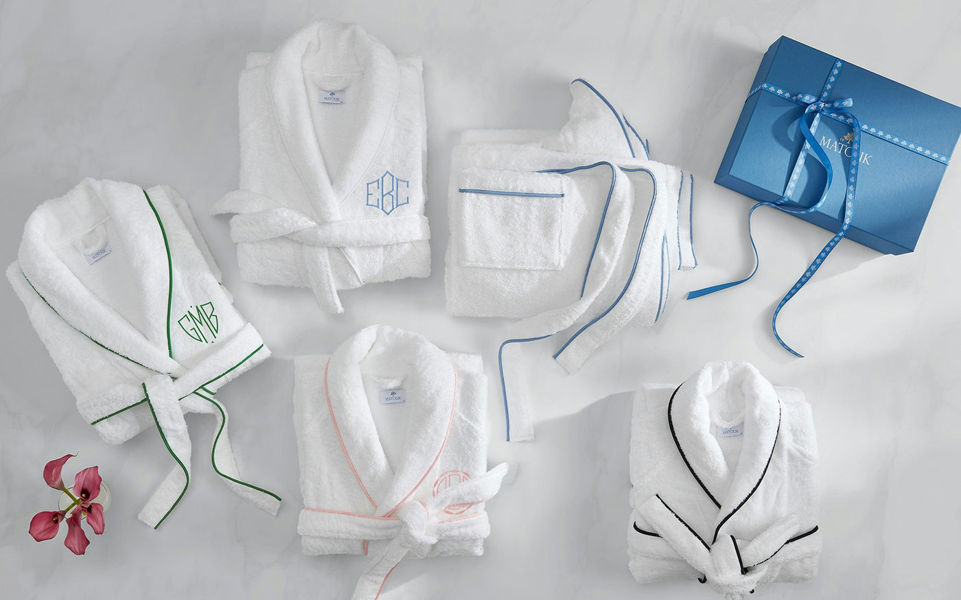 Close-up overhead view of multiple white Matouk robes with varied piping colors on marble surface.