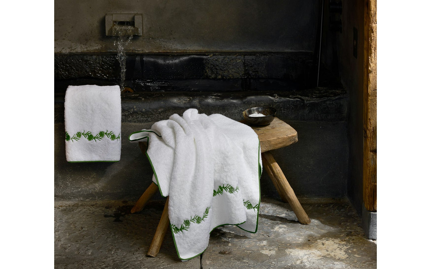 Close-up of Matouk Daphne tub mat draped over wooden stool with green floral embroidered border.