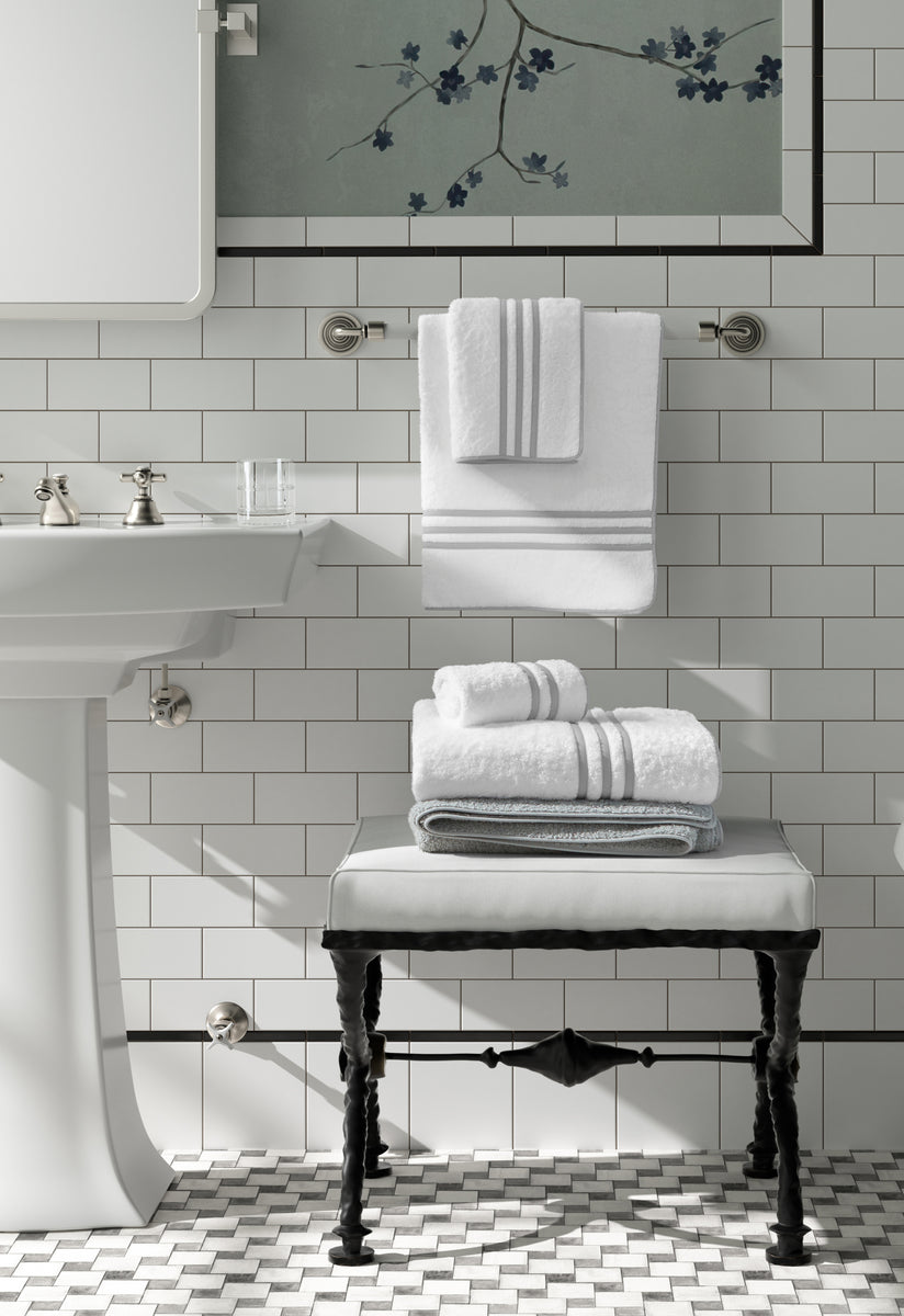 Matouk Newport wash cloths are neatly folded on a stool and hanging on a towel bar in a bath with white subway tile walls and a pedestal sink.