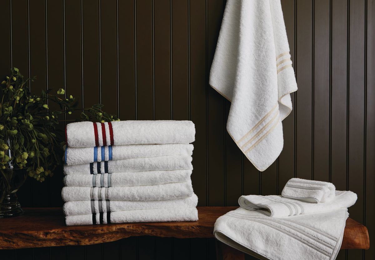 A Matouk Newport Framed Tub Mat is placed on a wooden bench, with additional white mats hanging and folded nearby, all set against a dark paneled bathroom wall.