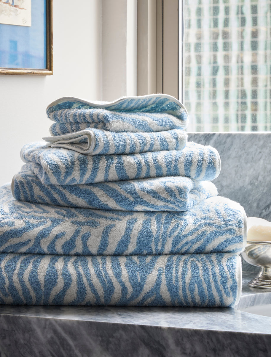 A stack of Matouk Regine wash cloths with a blue and white zebra jacquard design is neatly arranged on a marble countertop by the window.