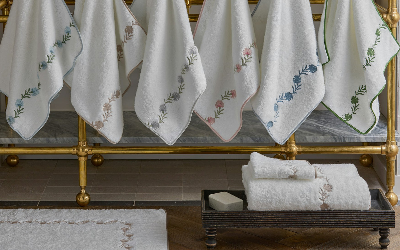 Folded white bath towels on a tray beside hanging embroidered white linens on a brass rack.