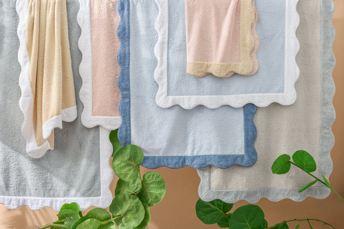 Several Neptune | Pool Towels by Matouk in pastel colors, featuring scalloped edges, hang in overlapping layers above green leafy plants against a beige background.