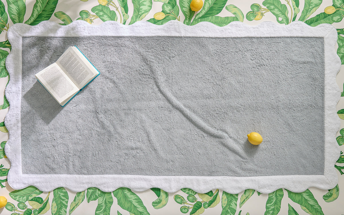The Neptune Pool Towel by Matouk, featuring scalloped edges in gray, is displayed on a leafy patterned surface with an open book and two lemons resting on top.