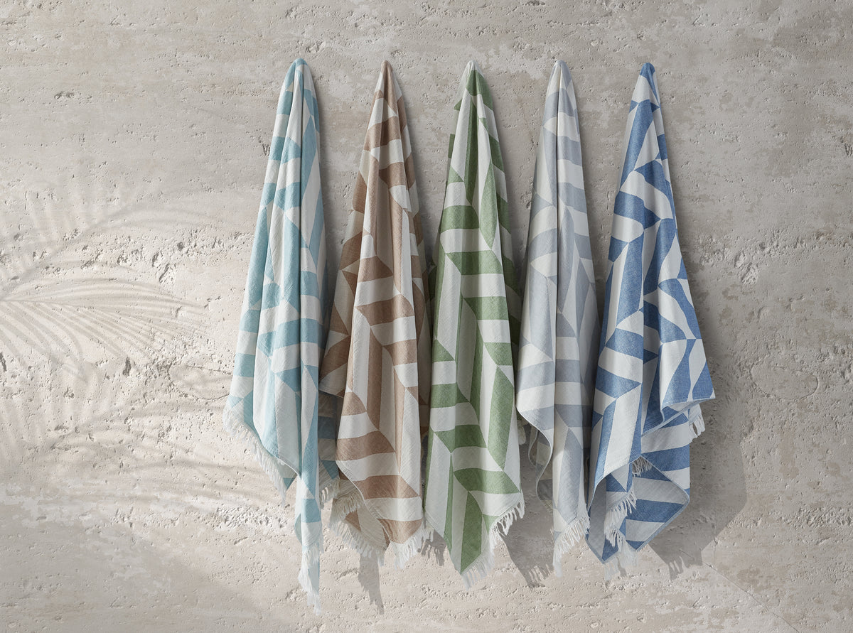 Five Paros Beach Towels by Matouk in pastel stripes—light blue, brown, green, gray, and blue—hang on hooks against a textured beige wall, highlighting the elegant simplicity of classic Turkish bath design.