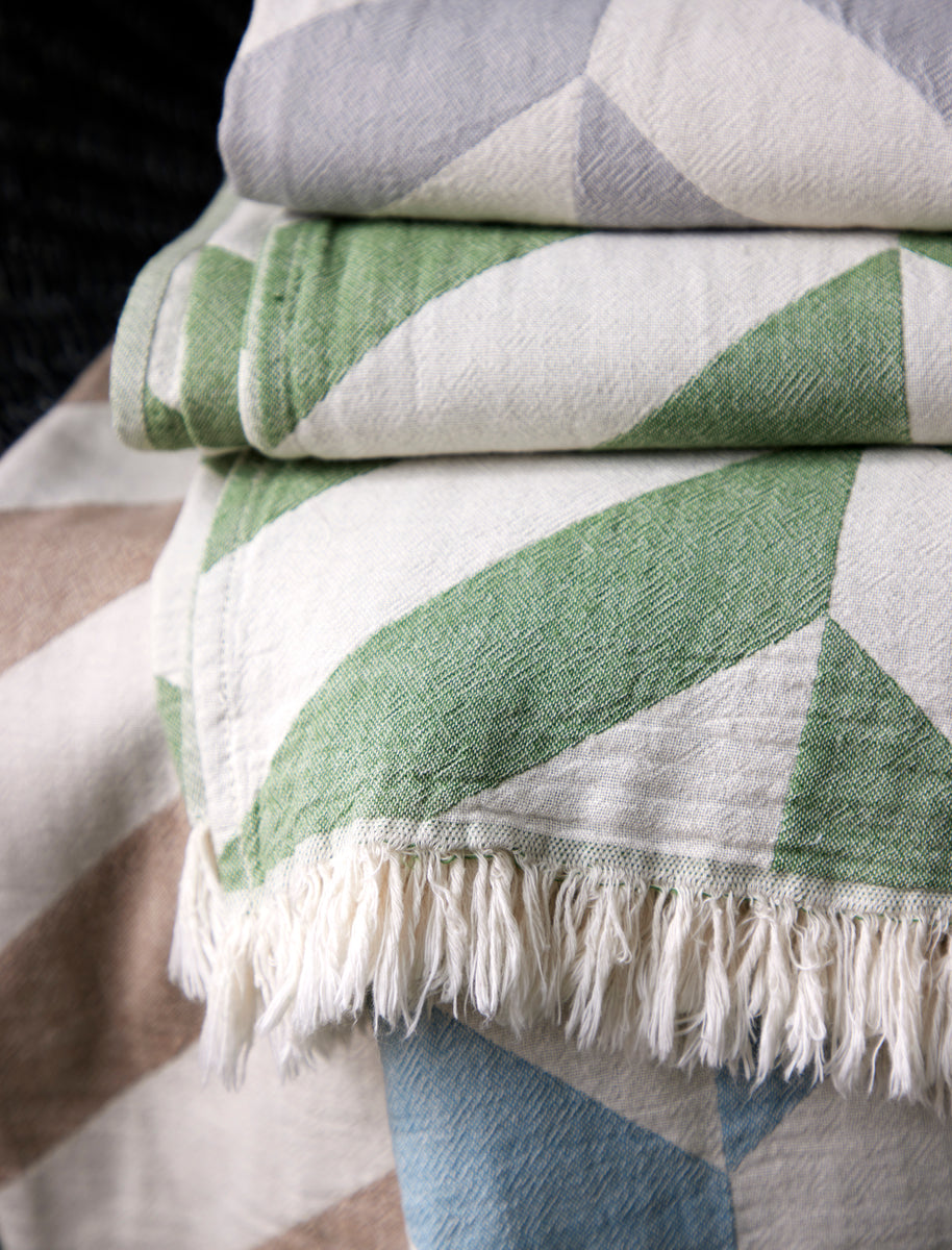 A close-up of Matouk’s Paros Beach Towel, featuring folded woven fabric with geometric patterns in green, blue, gray, and beige and fringe on one edge—evoking the look of a classic Turkish bath towel.