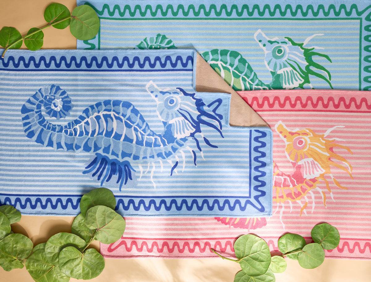 Three Matouk Seahorse Beach Towels in blue, pink, and green are displayed on a beige surface with green leaves scattered around them.