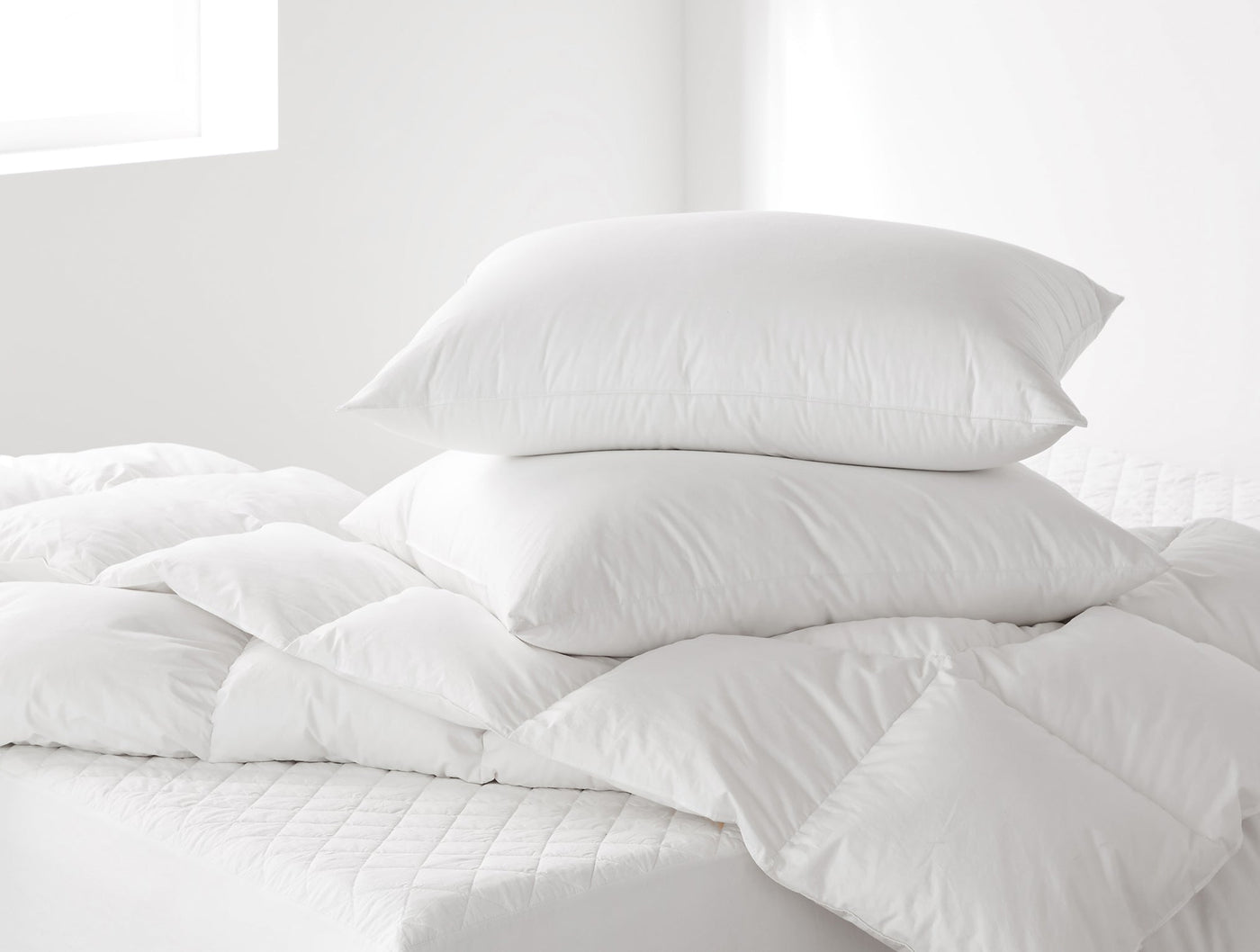 Two white down pillow inserts stacked on a quilted white comforter in a bright bedroom setting.