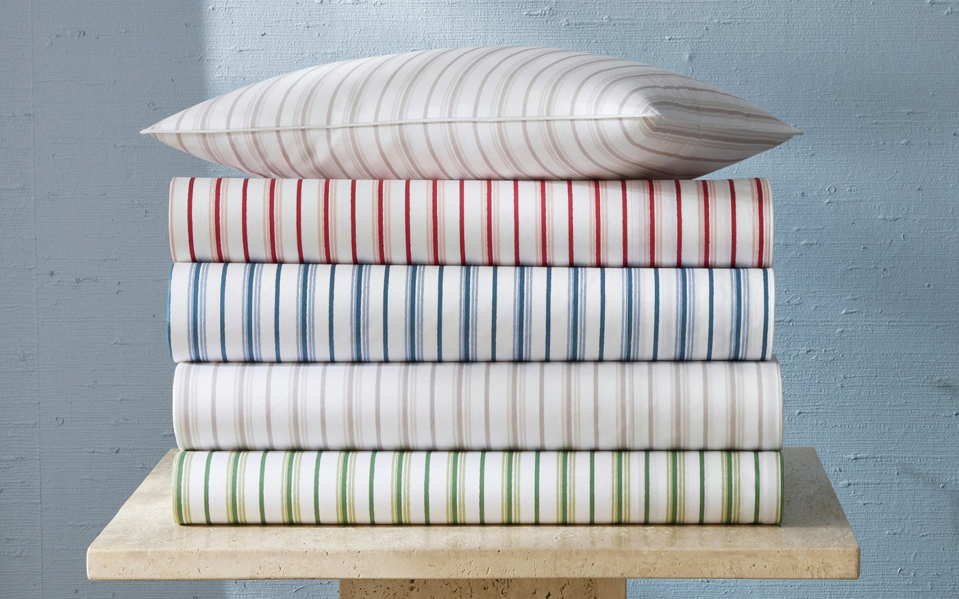 Stack of folded Matouk Calypso fitted sheets in four colorways on a stone surface.