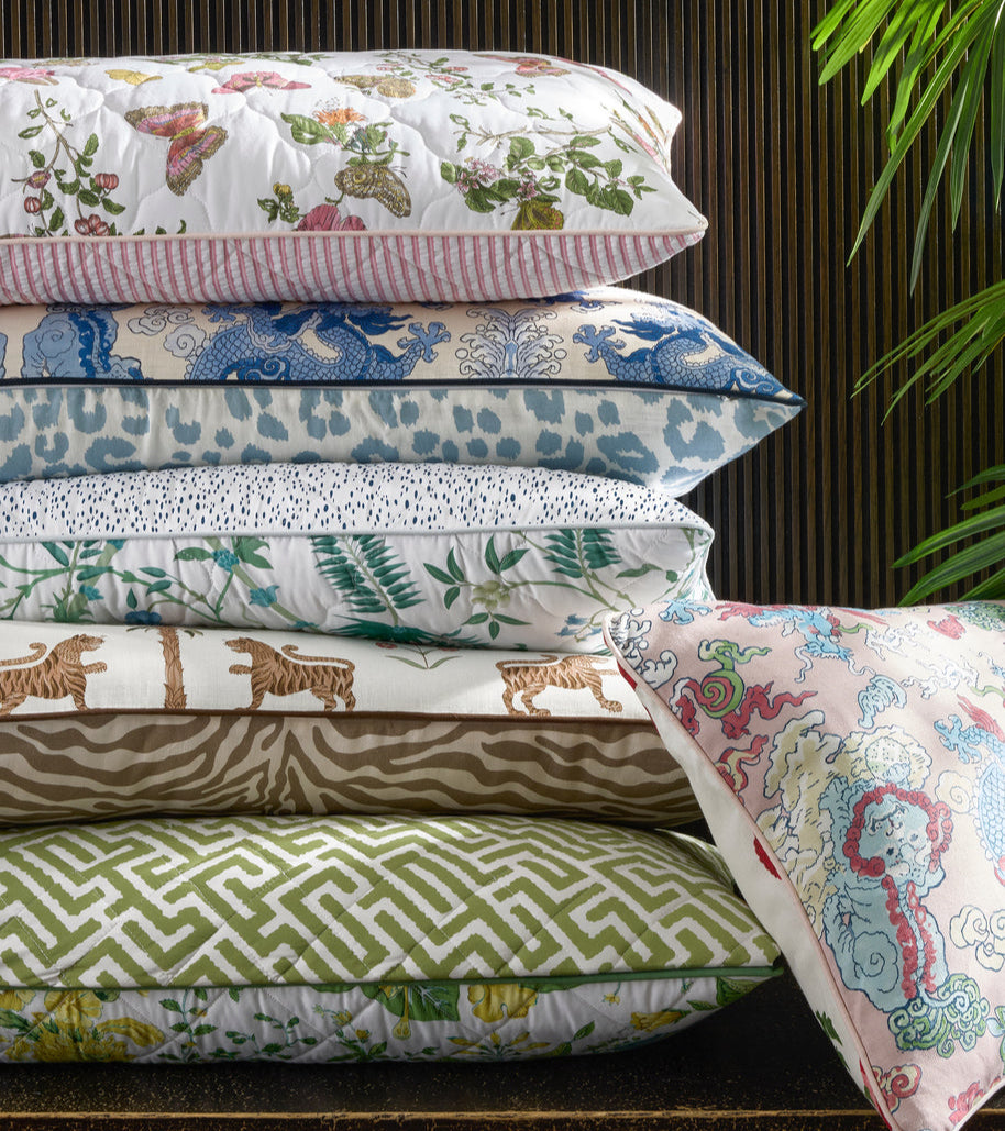 A stack of five decorative pillows with colorful patterns sits beside a sixth customizable Reversible Quilted Lumbar Pillow Cover by Matouk, with a green plant partially visible in the top right corner.