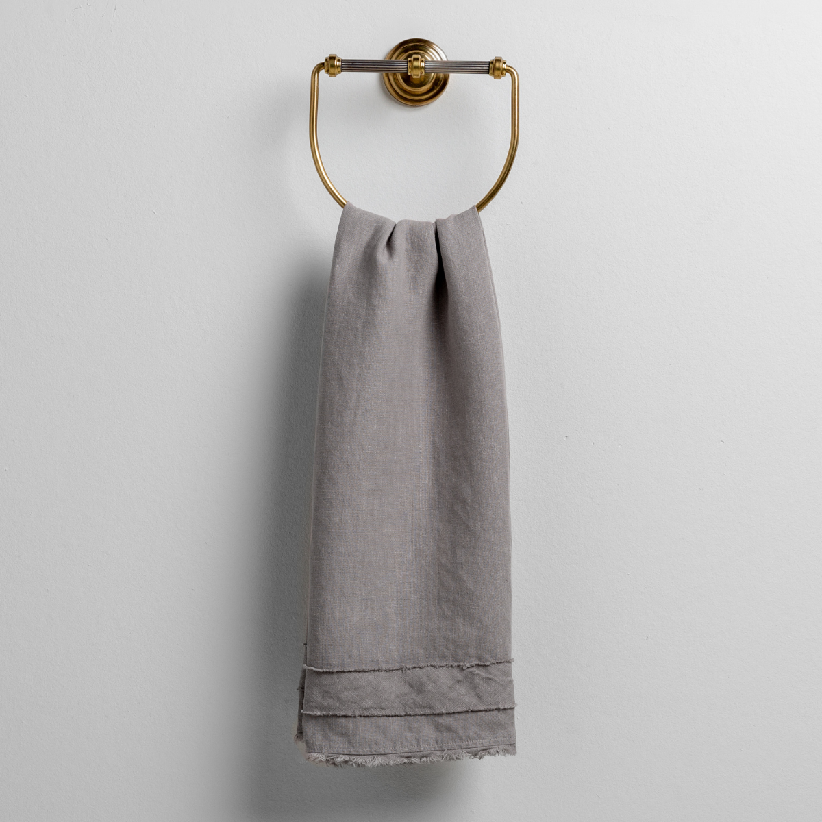 Fog linen guest towel draped over a brass ring wall mount, showing frayed hem detail.