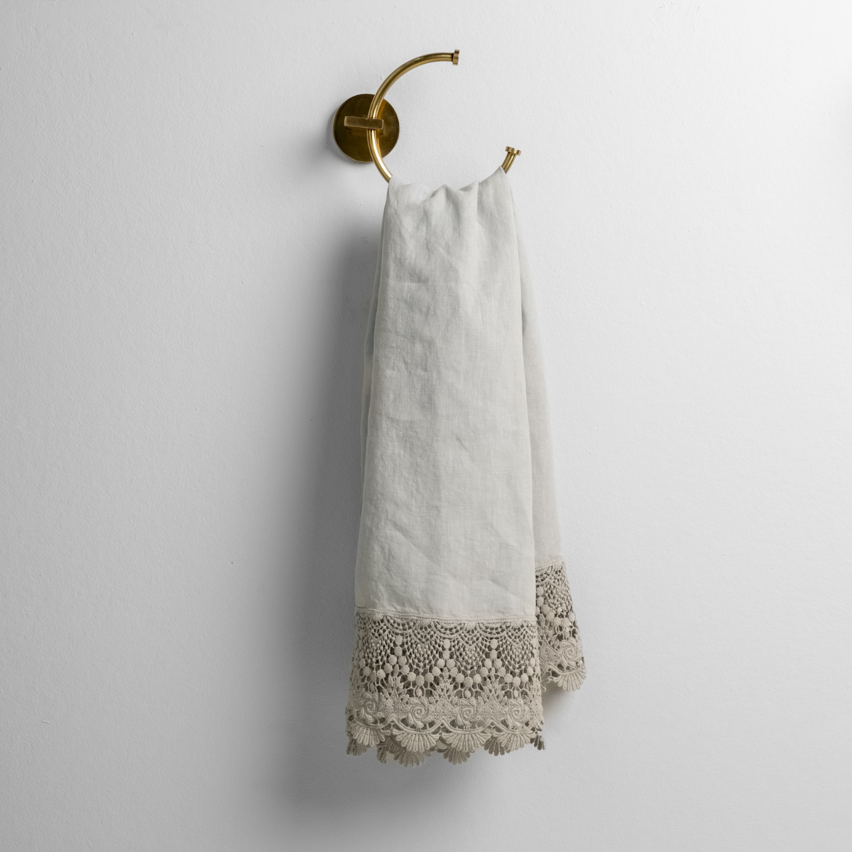 Cloud linen guest towel with wide crochet lace trim, hung on brass ring in bathroom.