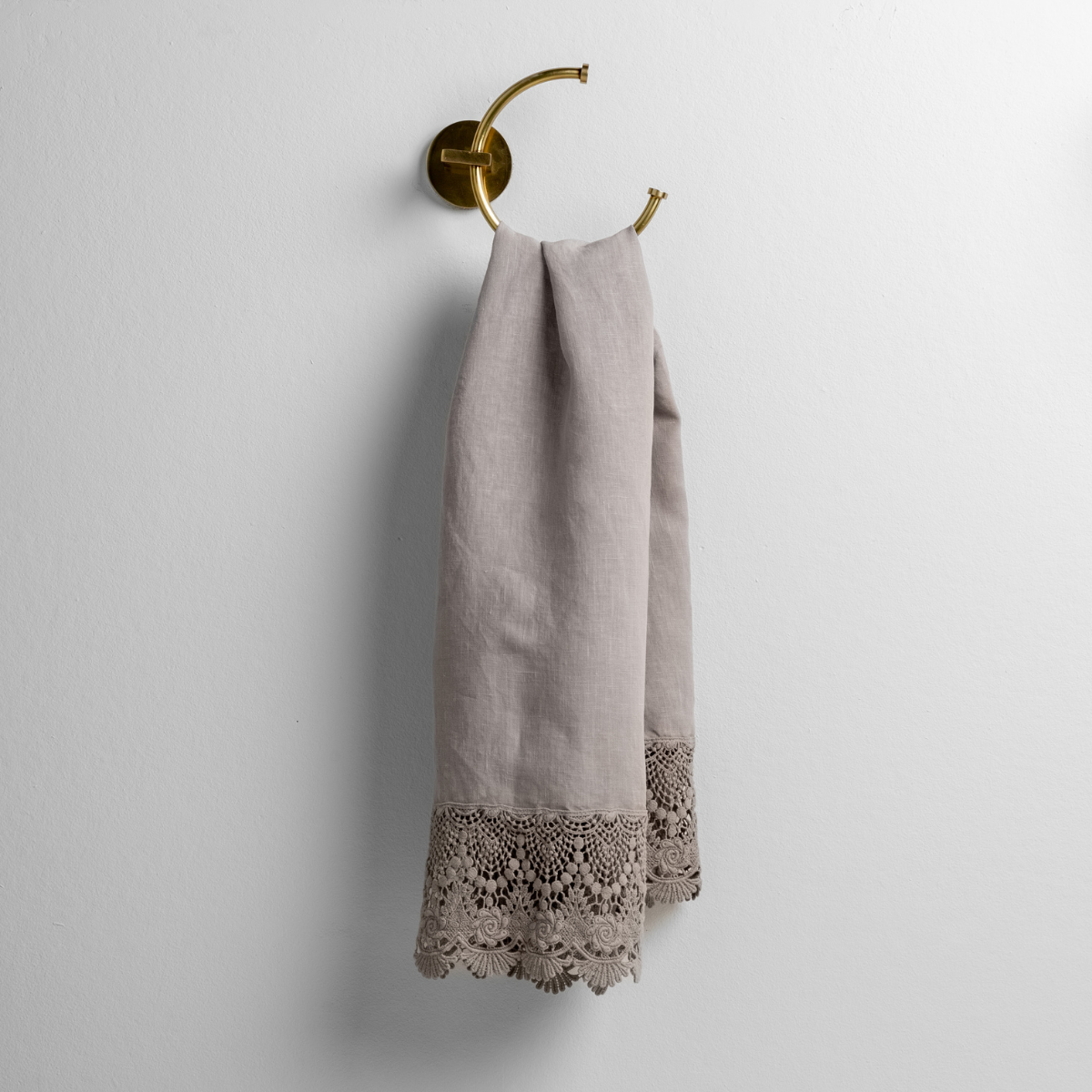 Fog guest towel hanging on brass ring hook, showing intricate lace trim detail at hem.