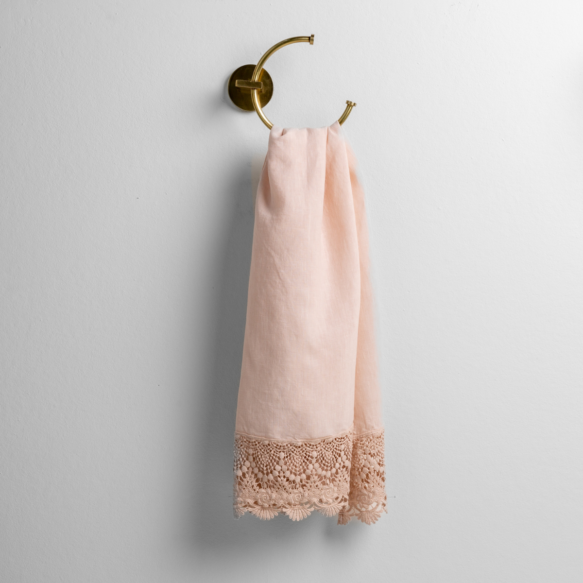 Pearl guest towel with scalloped lace trim, hung on brass ring hook against white wall.