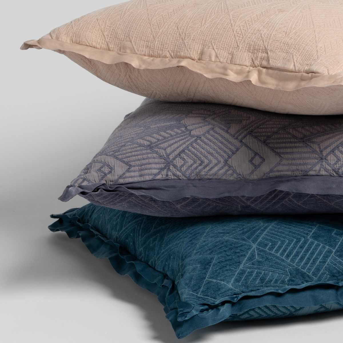 Three stacked Bella Notte Trettino throw pillows in taupe, slate, and teal with geometric jacquard pattern.