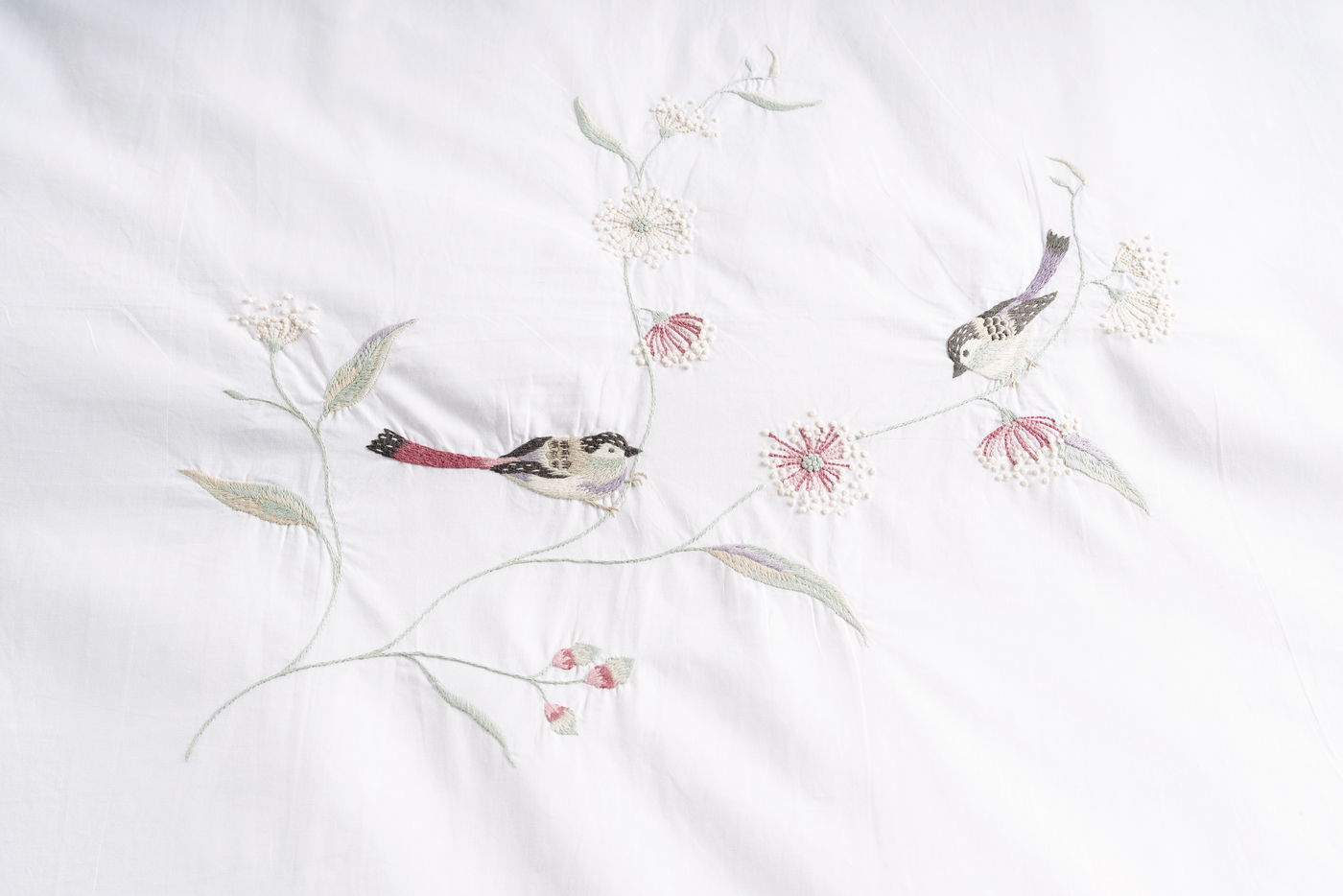 Close-up of hand-embroidered sparrows and red wildflowers on white cotton bedding.