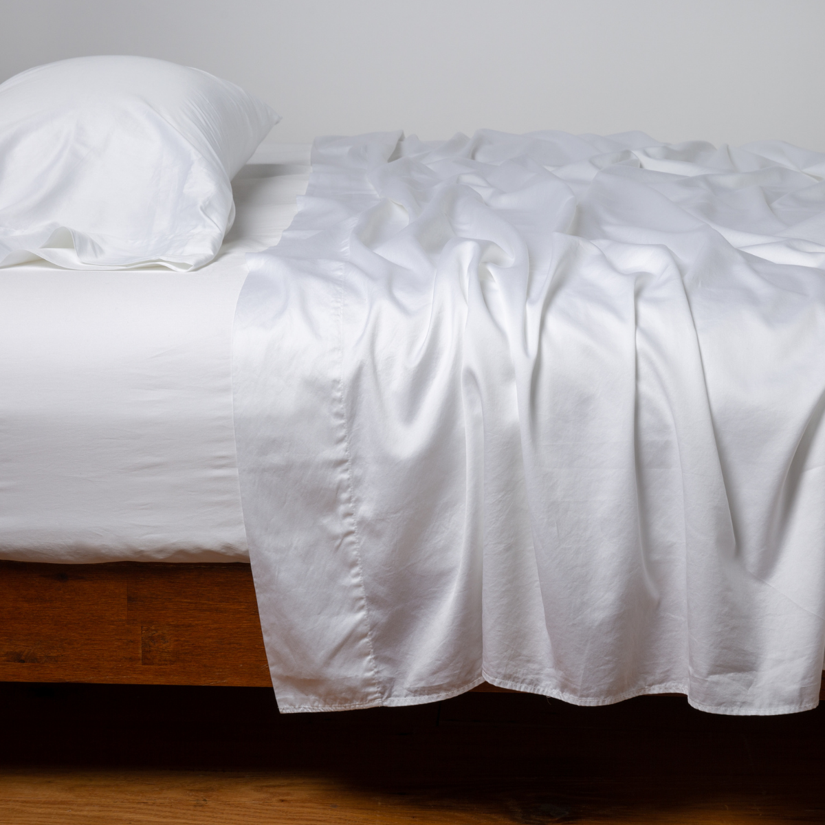White flat sheet draped loosely over a wood-frame bed with a matching pillow.