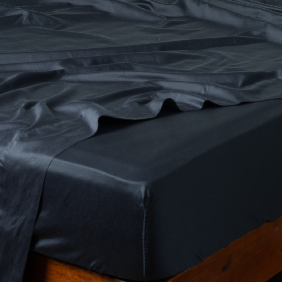 Corner detail of Bella Notte Bria fitted sheet in Slate, showing lustrous satin drape on a wood bed frame.