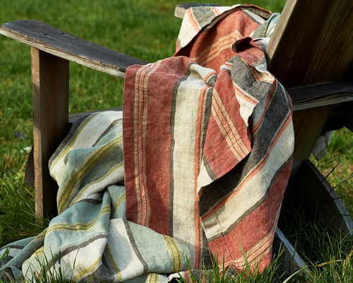 Rust, sage, and charcoal striped linen blanket draped over a weathered wood chair outdoors.