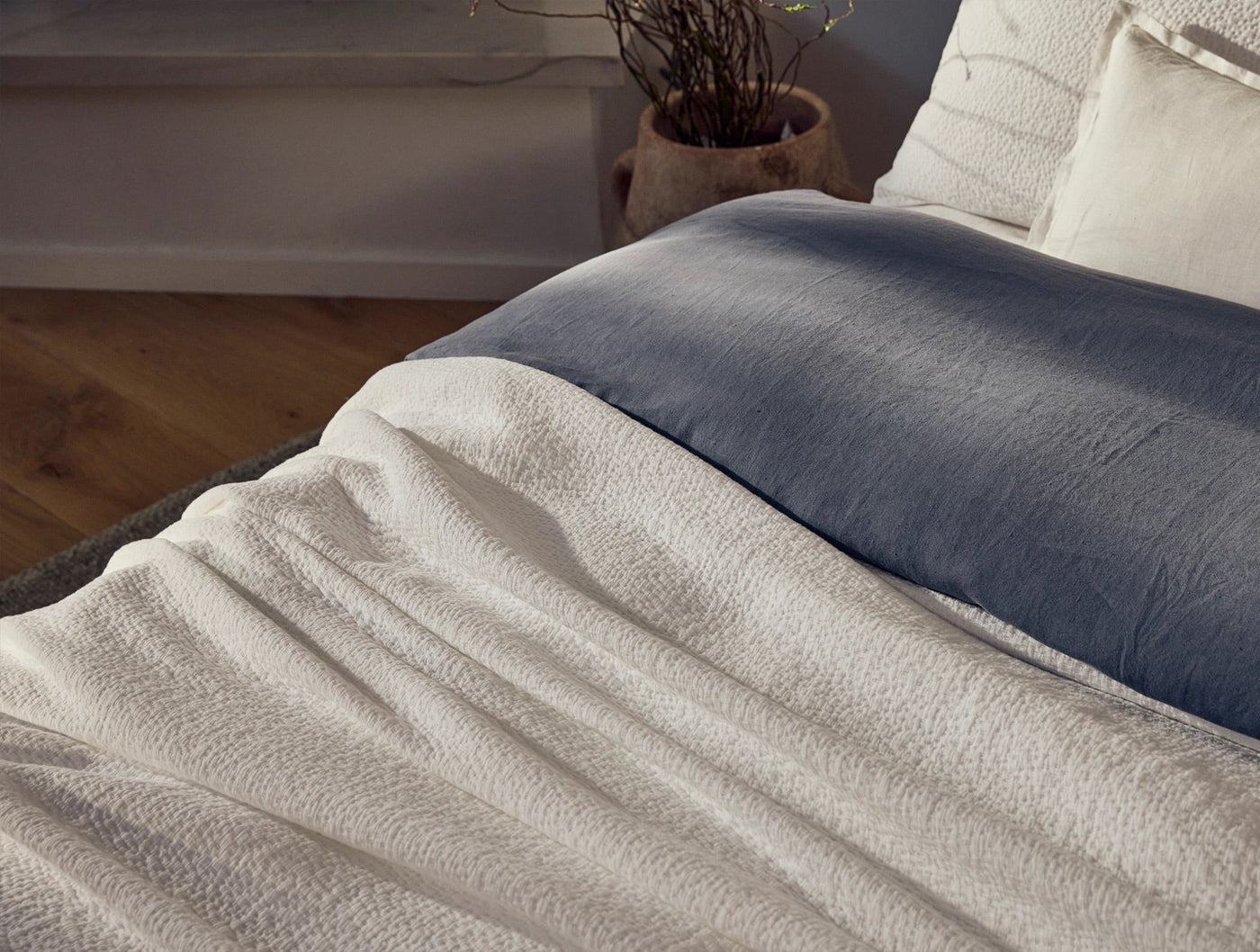 Close-up of ivory matelassé blanket draped over bed with slate blue linen layer beneath.