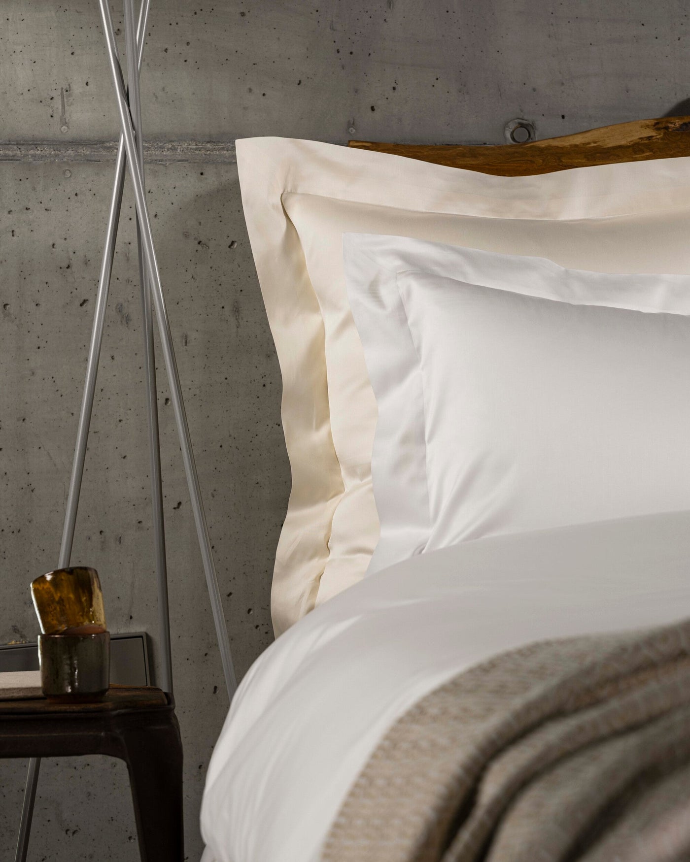 Close-up of ivory and white cotton pillowcases with trim detail against concrete wall.